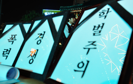 입시경쟁교육에 희생된 학생들을 위한 추모촛불문화제 '죽음의 교육을 멈춰라'에서 자유발언 중인 학생 앞으로 폭력과 경쟁, 학벌주의의 영정이 보이고 있다.